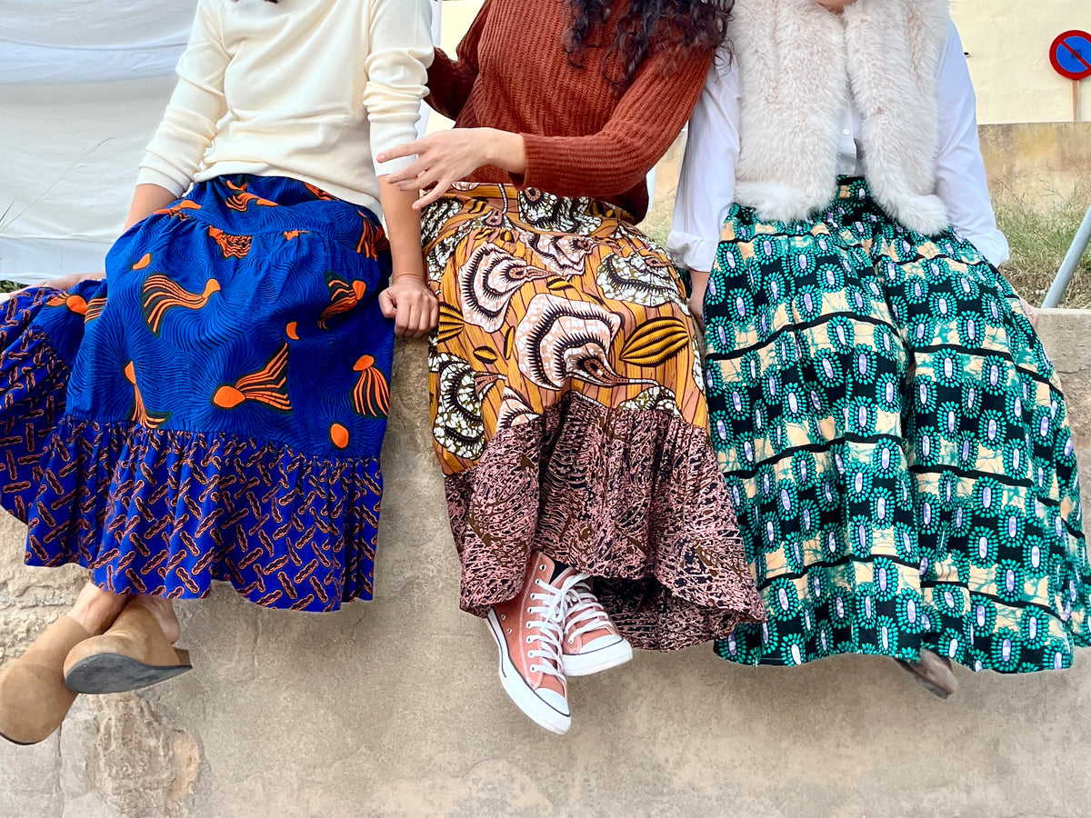 Tres mujeres sentadas luciendo faldas largas usawa con estampados africanos, azul, marrón y turquesa.